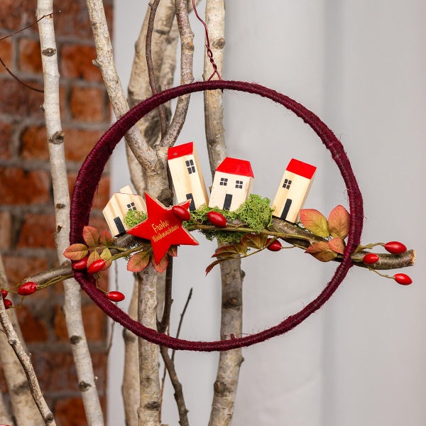 Weihnachtlicher Dekoring aus Metall mit umwickeltem Garn, dekoriert mit Holzhausfiguren, einem Stern mit Aufschrift und künstlichen Hagebutten.