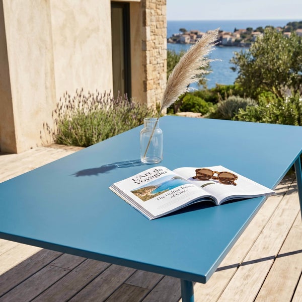 Blauer Gartentisch aus Metall auf einer Holzterrasse mit Meerblick, dekoriert mit Vase, Magazin und Sonnenbrille.