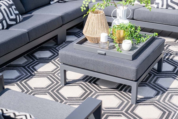 Moderne Gartenlounge mit grauen Polstern, quadratischem Hocker als Tisch mit Dekotablett und geometrisch gemustertem Outdoorteppich auf einer Terrasse.