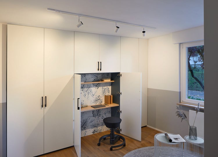 Moderner weißer Einbauschrank mit integriertem Schreibtisch für Homeoffice, Holzregalen und Schienenbeleuchtung in einem hellen Zimmer.