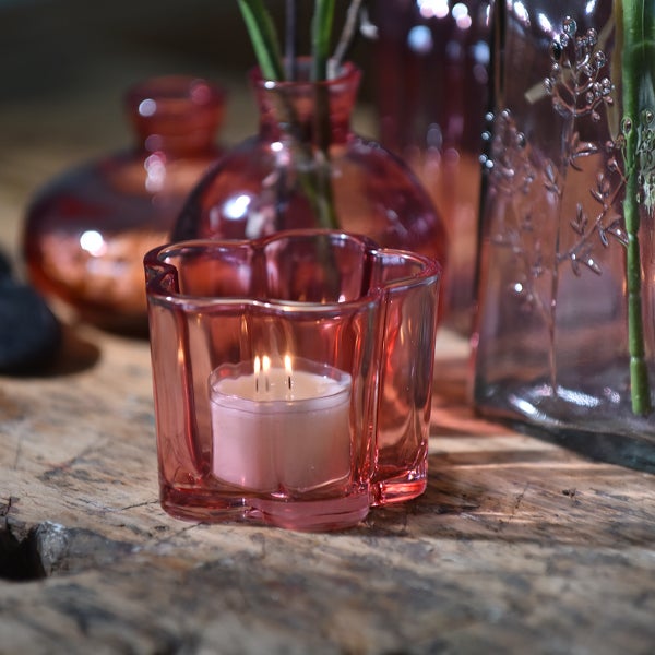 Windlicht aus rosafarbenem Glas in Blumenform mit brennendem Teelicht auf einem Holztisch.