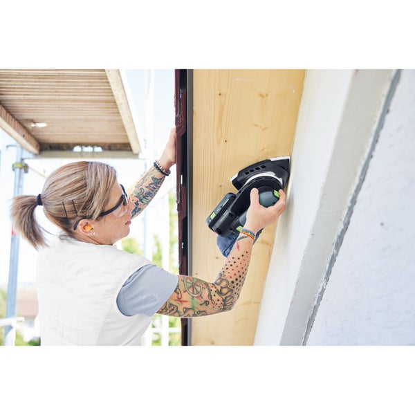Handwerkerin schleift ein Holzbrett unter einem Dachüberstand mit einem Akku-Multischleifer und trägt dabei eine Schutzbrille und Gehörschutz.