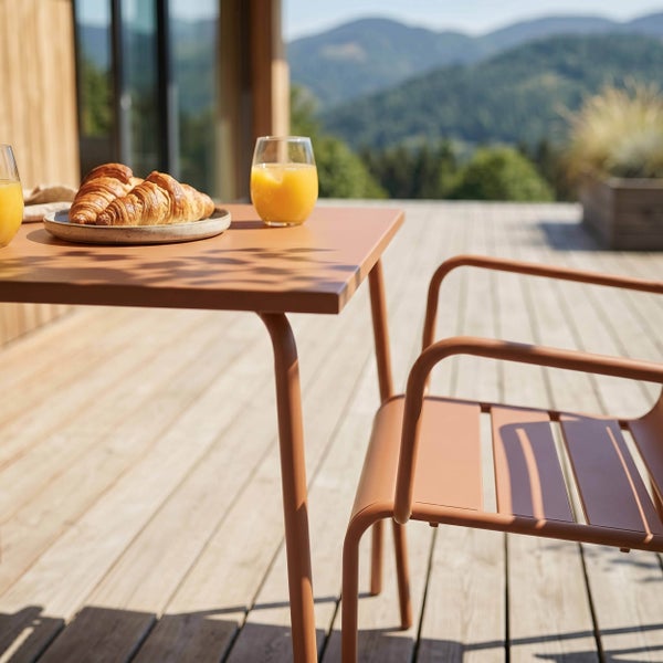 Moderner Gartentisch und Gartenstuhl aus Metall in Terracotta auf einer hellen Holzterrasse mit Frühstück und Bergpanorama.