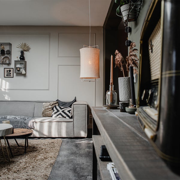 Modernes Wohnzimmer mit grauem Sofa, Teppich, Holztisch und einer zylindrischen Textilpendelleuchte vor einer Wand mit Zierleisten.