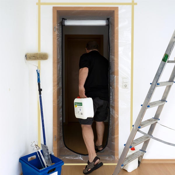 Ein Mann geht durch eine Staubschutztür mit Reißverschluss in einem hellen Raum mit Malerwerkzeugen wie Farbwalze und Leiter.