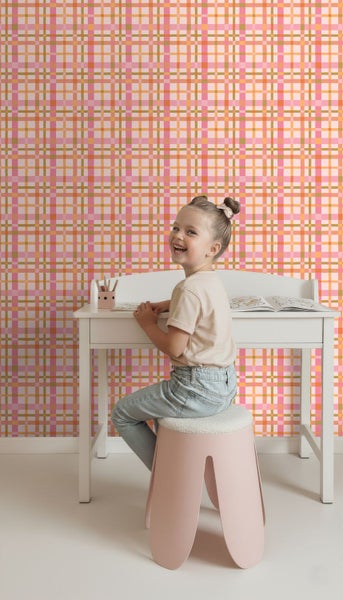 Vliestapete mit Karomuster in Rosa und Orange hinter einem weißen Kinderschreibtisch in einem hellen Kinderzimmer.