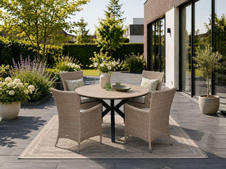 Runder Gartentisch aus Holz mit vier Korbstühlen auf einem Outdoor-Teppich auf einer Terrasse, im Hintergrund ein grüner Garten.
