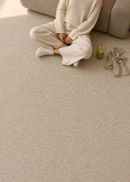 Eine Person sitzt im Schneidersitz auf einem hellen, strukturierten Teppichboden in einem hellen Wohnzimmer neben einem Sofa.