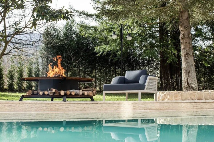 Gartenlounge mit Feuertisch und Sessel am Poolrand unter Bäumen mit brennendem Feuer im Feuertisch