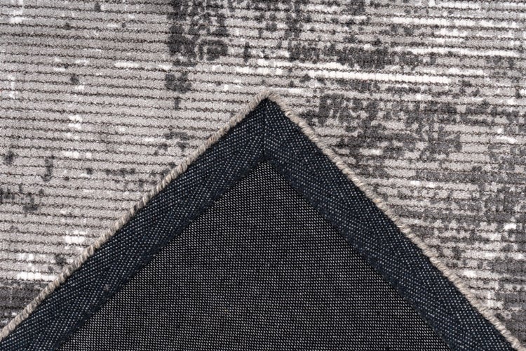 Nahaufnahme eines Kurzflor-Teppichs mit grau-weißem Vintage-Muster, umgeknickte Ecke zeigt die strukturierte Rückseite und den gekettelten Rand.