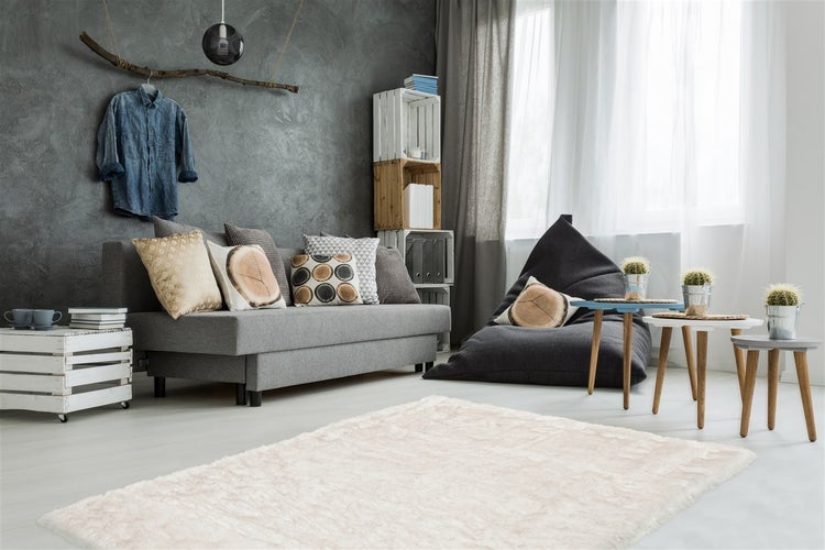 Modernes Wohnzimmer mit grauem Sofa, Sitzsack, Holzkistenregal, Beistelltischen und flauschigem Teppich vor einer strukturierten Wand.