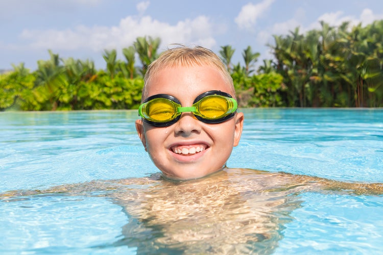 Ein Kind mit Schwimmbrille im Wasser eines Pools vor einer Kulisse aus tropischen Pflanzen.