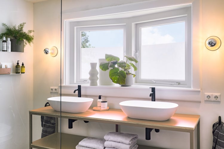 Modernes helles Badezimmer mit zwei ovalen weißen Aufsatzwaschbecken, schwarzen Armaturen und Holzkonsole unter einem Fenster.