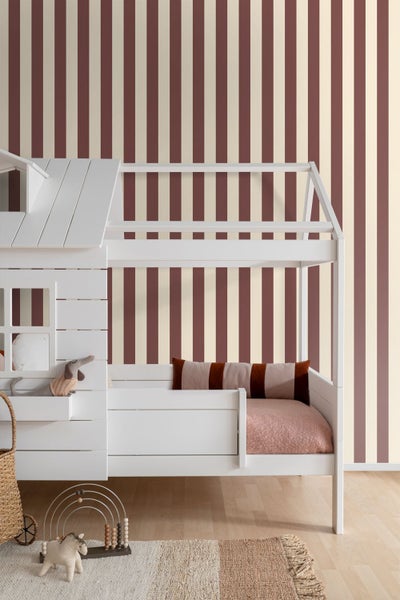Ein helles Kinderzimmer mit einer vertikal gestreiften Tapete in Bordeaux und Creme hinter einem weißen Hausbett aus Holz.