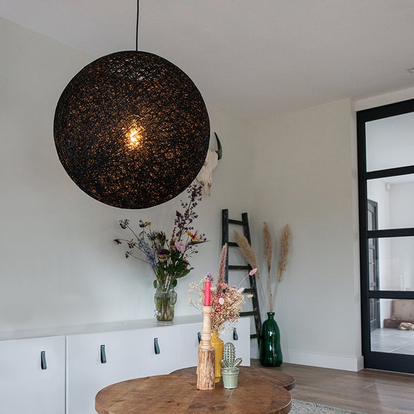 Schwarze, kugelförmige Pendelleuchte aus Geflecht in einem hellen Wohnzimmer mit weißem Sideboard und Holzmöbeln.