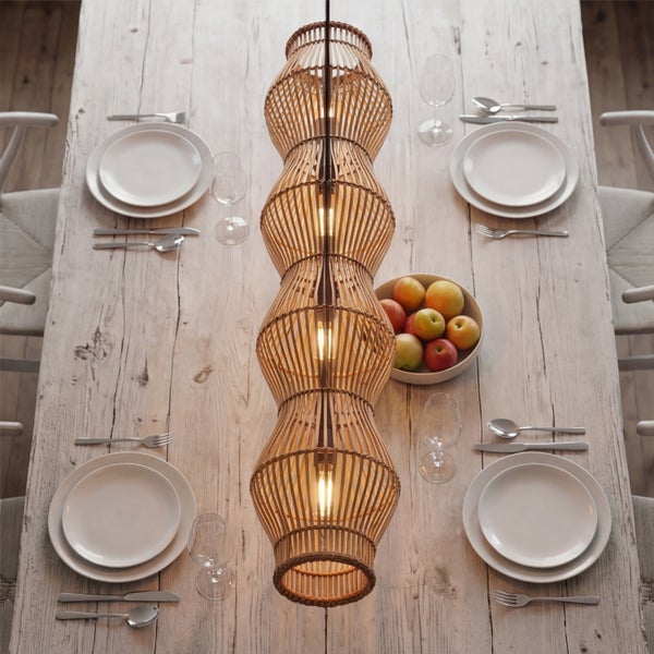Längliche Pendelleuchte aus Holzgeflecht über einem rustikal gedeckten Esstisch aus hellem Holz mit Tellern und Besteck.