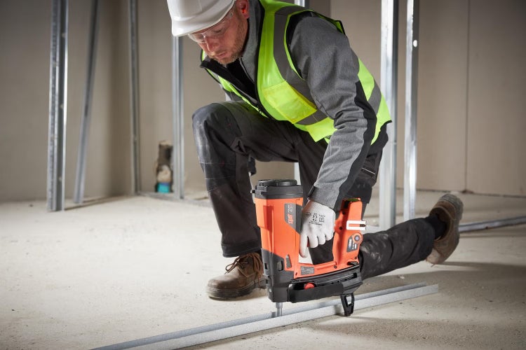 Handwerker mit Schutzhelm und Warnweste befestigt ein Metallprofil im Trockenbau mit einem Gasnagler auf einem Betonboden.