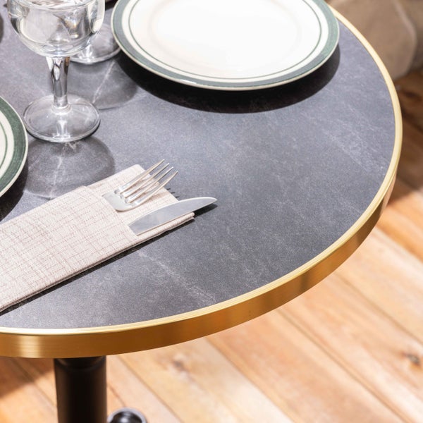 Runder Bistrotisch mit dunkler Platte in Steinoptik und goldfarbenem Rand, gedeckt mit Teller, Glas und Besteck.