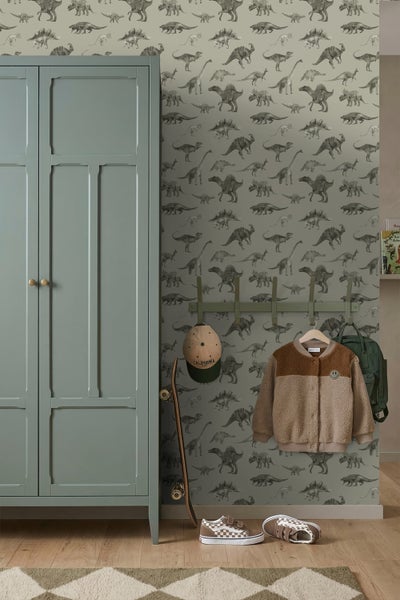 Vliestapete mit Dinosaurier-Motiv in einem organisierten Kinderzimmer neben einem grünen Kleiderschrank und einer Garderobe mit Kinderjacke.