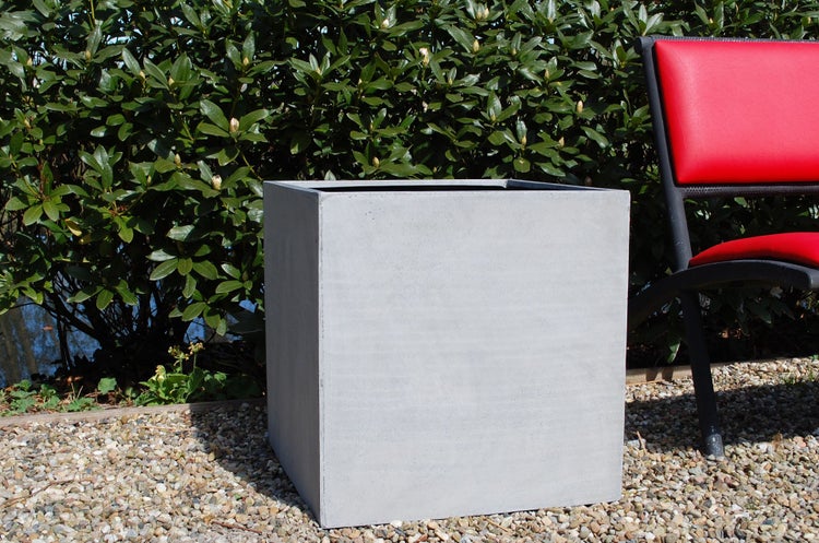 Quadratischer grauer Pflanzkübel in Betonoptik im Garten auf Kies vor einer grünen Hecke.