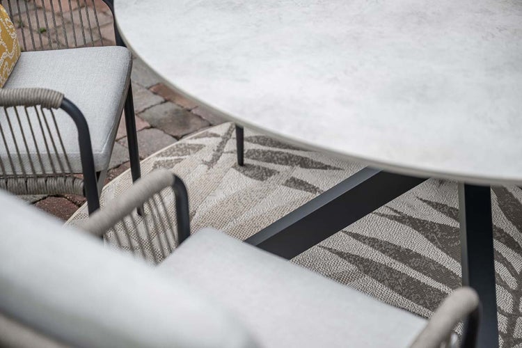 Moderne Garten-Essgruppe mit rundem Tisch in Steinoptik, Seilgeflecht-Stühlen mit Kissen und einem gemusterten Außenteppich auf einer Terrasse.
