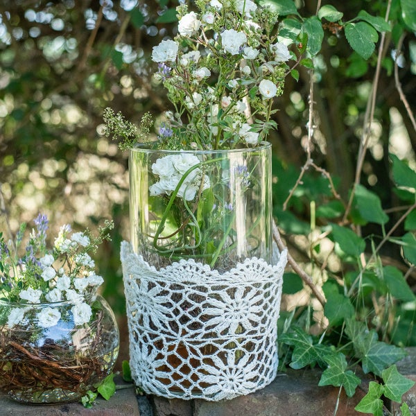 Zwei Glasvasen mit weißen Blumen auf einer Steinmauer, eine mit Häkelspitze verziert.