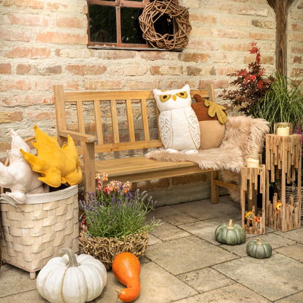 Herbstliche Terrassengestaltung mit Holzbank, Kürbissen, dekorativen Kissen, einer Weidenkorb-Pflanzschale und Windlichtern vor einer Backsteinwand.