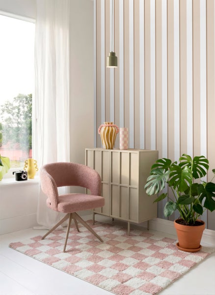 Modernes helles Zimmer mit vertikal gestreifter Tapete, einem rosa Sessel, einem beigen Sideboard, einem Schachbrettteppich und einer Monstera-Pflanze.
