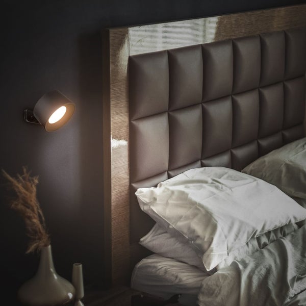 Wandmontierte Leselampe neben einem Bett mit gepolstertem Kopfteil in einem Schlafzimmer.