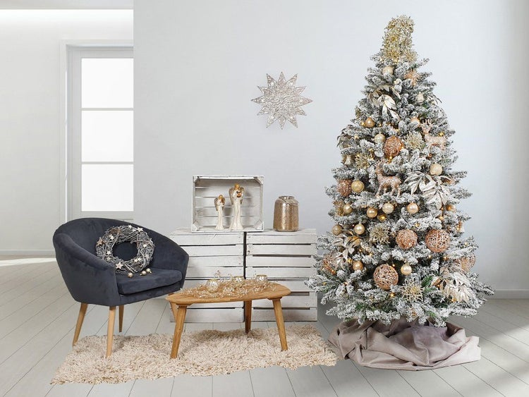Festlich geschmückter, künstlicher Weihnachtsbaum mit Schneeoptik, dazu ein Sessel, Holzkisten, Tisch und weihnachtliche Dekoration im Wohnraum.