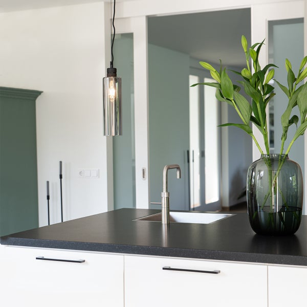 Moderne Kücheninsel mit dunkler Arbeitsplatte, Edelstahl-Armatur und Spüle, zylindrischer Pendelleuchte aus Glas und Vase mit grünen Lilien.