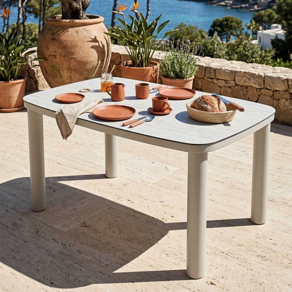 Rechteckiger Gartentisch mit abgerundeten Ecken und dicken Beinen, gedeckt mit Terracotta-Geschirr auf einer sonnigen Terrasse mit Meerblick.