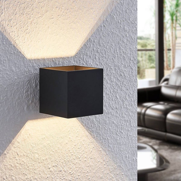Moderne schwarze Wandleuchte in Würfelform an einer strukturierten Wand mit Lichtstrahlen nach oben und unten, im Hintergrund ein Sofa aus Leder.
