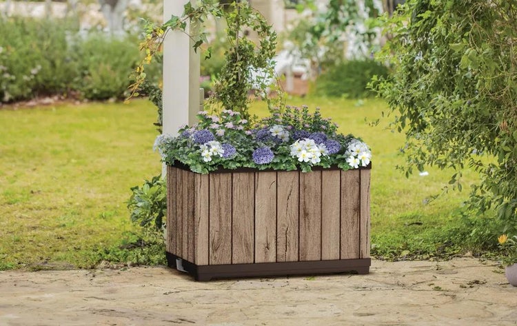 Rechteckiger Pflanzkasten in Holzoptik mit vertikalen Lamellen, bepflanzt mit Blumen, auf einer Steinterrasse in einer hellen Gartenumgebung.