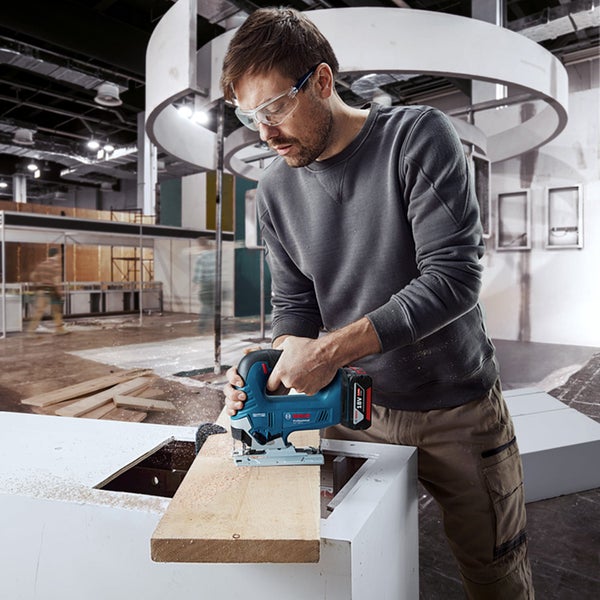 Ein Handwerker sägt mit einer blauen Bosch Akku-Stichsäge ein Holzbrett in einer hellen Werkstattumgebung.