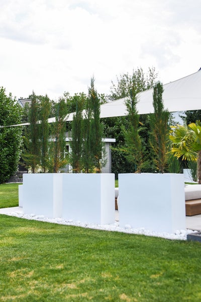 Moderne Gartengestaltung mit rechteckigen Pflanzkübeln, schmalen Nadelbäumen, einem Sonnensegel und einer gepflegten Rasenfläche.
