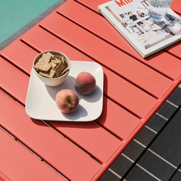 Roter Gartentisch aus Metalllamellen mit Pfirsichen, Crackern, einer Zeitschrift und einem Glas Wasser in hellem Sonnenlicht.