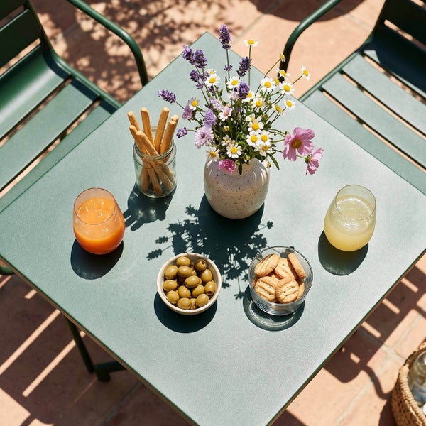 Grünes Gartenmöbel-Set aus Metall bestehend aus Tisch und Stühlen auf einer Terrasse mit Blumen, Getränken und Snacks.