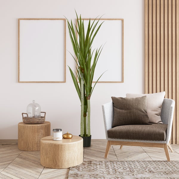 Heller Raum mit zwei Holzbilderrahmen, einer hohen Kunstpflanze, einem Korbsessel mit Kissen und runden Holztischen auf Fischgrätparkett.