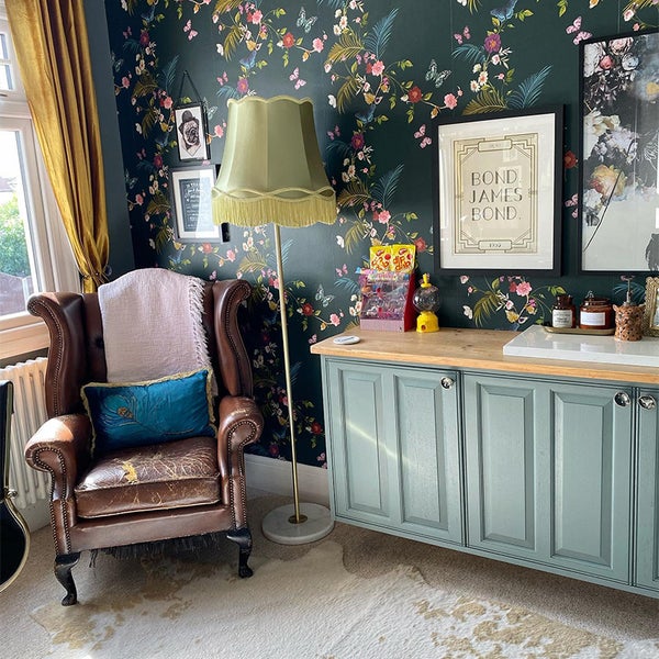 Wohnzimmer mit Ohrensessel, Stehlampe und Kommode vor einer floralen Tapete