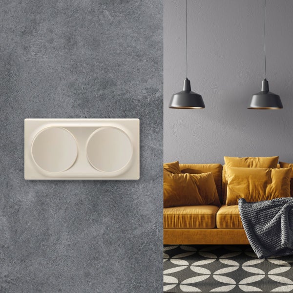 Doppelwippschalter auf einer grauen strukturierten Wand neben einem Wohnzimmer mit gelbem Sofa und zwei Pendelleuchten.
