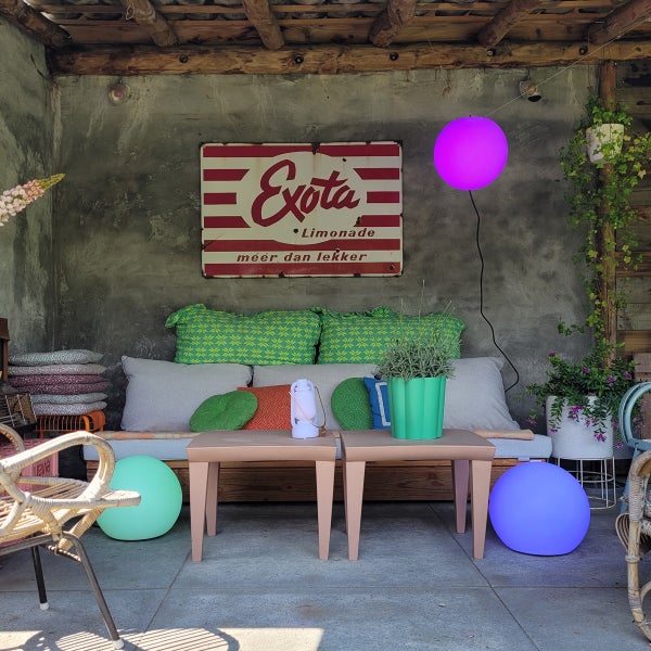 Terrasse mit Holzbank, bunten Kissen, niedrigen Beistelltischen und leuchtenden Kugellampen vor einer Betonwand.