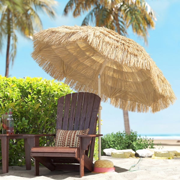 Sonnenschirm aus Naturbast über einem dunklen Adirondack-Gartensessel aus Holz mit gemustertem Kissen auf sandigem Untergrund am Strand.