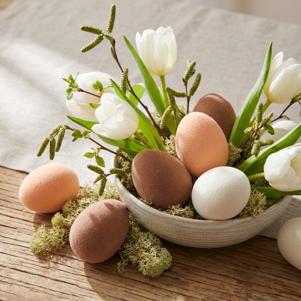 Osterdekoration mit beflockten Eiern in verschiedenen Brauntönen und weissen Tulpen in einer Keramikschale auf einem Holztisch.