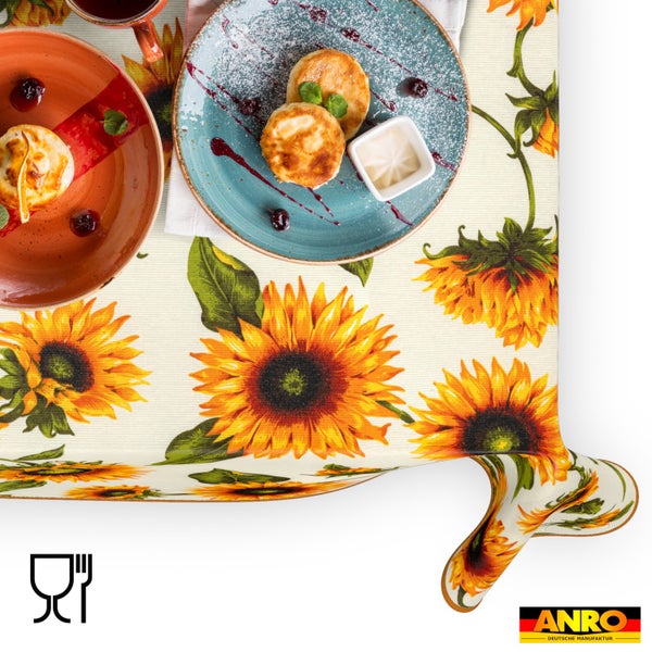 Tischdecke mit Sonnenblumen-Motiv, gedeckt mit Tellern und Gebäck. ANRO DEUTSCHE MANUFAKTUR Logo und Symbol für Lebensmittelechtheit.