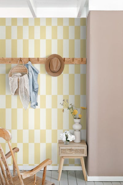 Wand mit gelb-weiß karierter Tapete, Holzgarderobe mit Korb und Jacke sowie kleinem Beistelltisch in einem hellen, organisierten Raum.