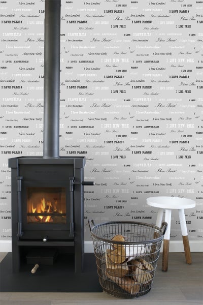 Raumansicht mit Kaminofen, Holzkorb mit Brennholz und Beistelltisch vor einer Tapete mit Städtenamen