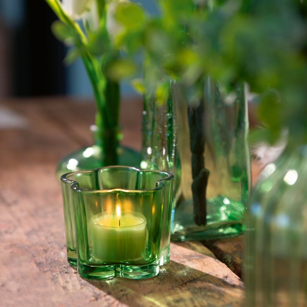 Grünes Teelichtglas in Blumenform mit brennender Kerze auf einem Holztisch, im Hintergrund grüne Vasen mit Pflanzen.