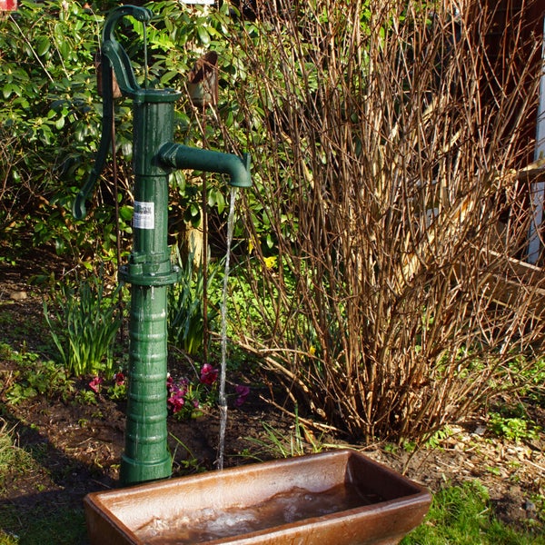 Eine historische Schwengelpumpe aus Metall mit fließendem Wasser über einem rechteckigen Becken in einem Garten.
