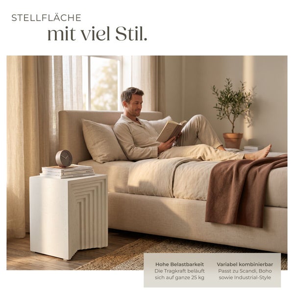 Mann liest auf einem Bett neben einem strukturierten, würfelförmigen Beistelltisch mit 25 Kilogramm Tragkraft in einem hellen Schlafzimmer.